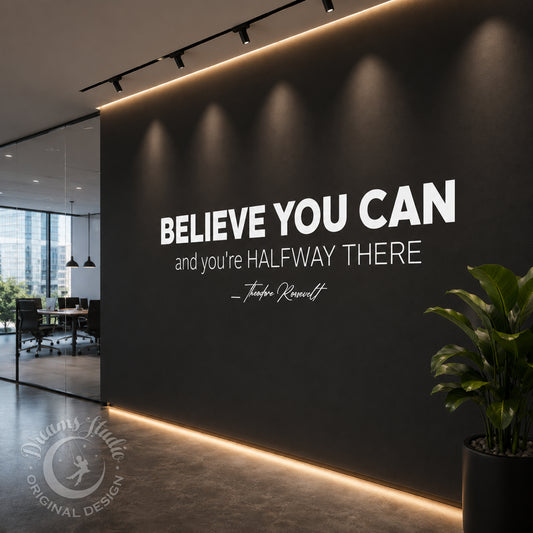 A modern office interior with a dark gray wall featuring the motivational quote 'BELIEVE YOU CAN and you're HALFWAY THERE — Tim Keller' in large white text. Track lighting casts soft beams onto the wall, and a potted plant sits in the lower right corner.