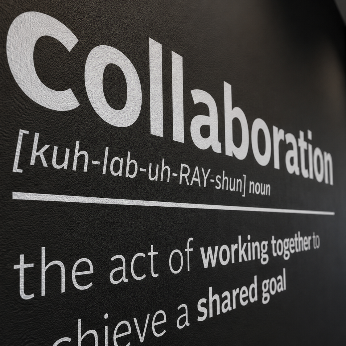 A close-up of a black wall or sign with large white text reading 'Collaboration' and a dictionary-style definition, plus the phrase 'the act of working together to achieve a shared goal.'.
